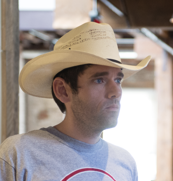 A person wearing a cowboy hat and a T-shirt stands indoors, looking thoughtfully to the side. The background is softly blurred, suggesting an industrial or rustic setting.